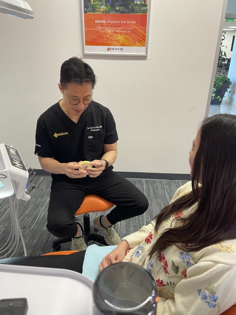 Dr. Kevin Kim, DDS, a dental implant specialist in a black uniform, sits with a patient in a Lakewood clinical setting, explaining an implant-retained overdenture using a yellow anatomical jaw model.